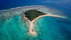 Vue aérienne d’un îlot corallien bordé de deux pointes de sable dans un lagon turquoise.