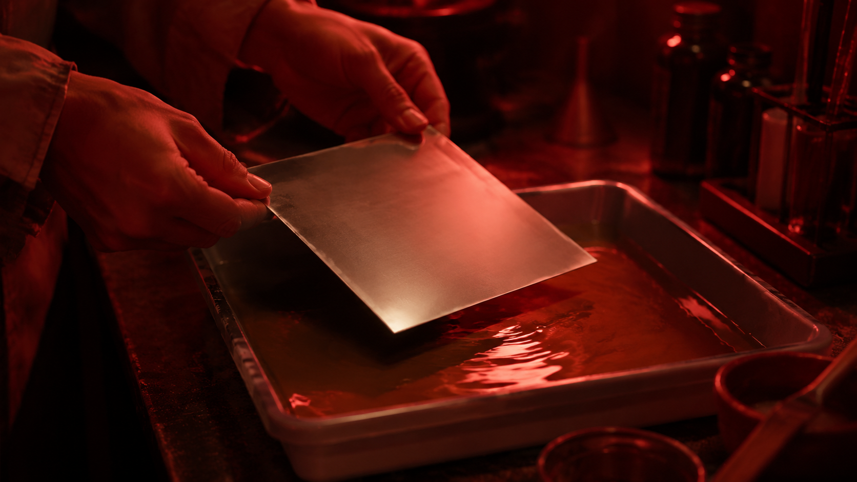 Mains anonymes tenant une plaque métallique vierge au-dessus d'un bac de révélateur, dans la lueur rouge d'un laboratoire.