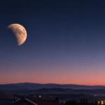 Une lune presque pleine partiellement assombrie par l'ombre de la Terre, basse au-dessus d'un horizon de collines et de toits à l'aube, ciel bleu nuit virant au rose.