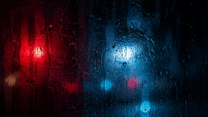 Vitre couverte de gouttes de pluie la nuit, halos de néons rouges et bleus flous derrière le verre.