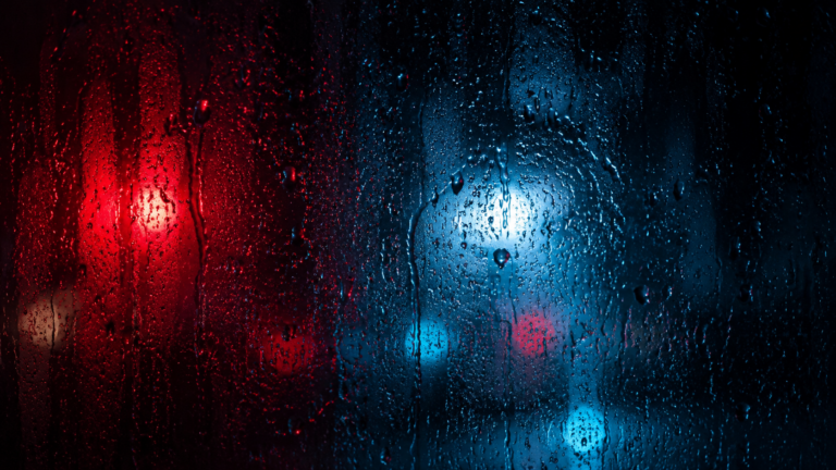 Vitre couverte de gouttes de pluie la nuit, halos de néons rouges et bleus flous derrière le verre.