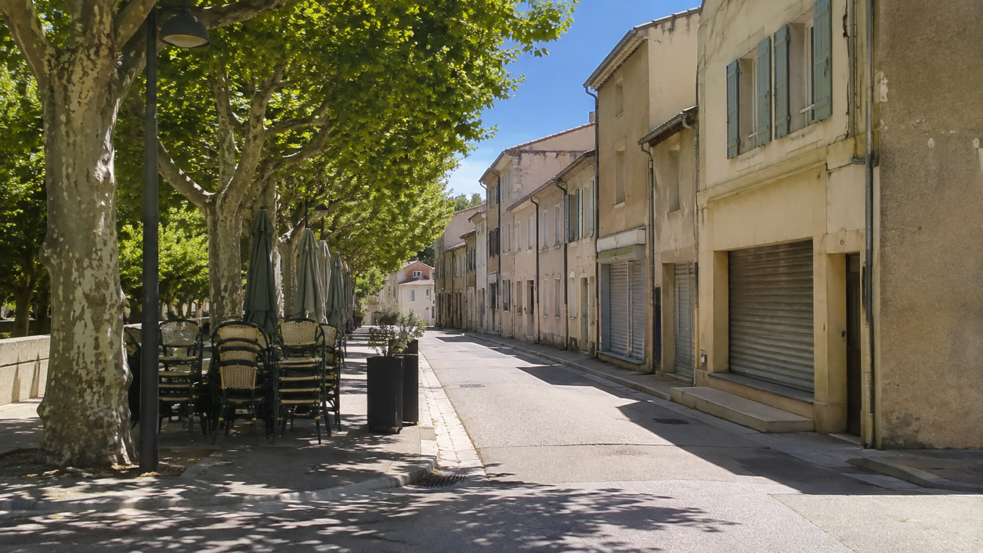 Rue de village française déserte à midi en août, rideaux de fer baissés sur les commerces et terrasse de café repliée sous les platanes