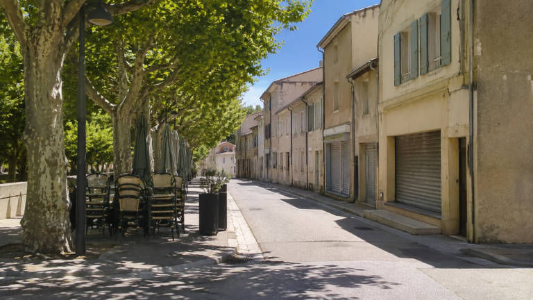 Rue de village française déserte à midi en août, rideaux de fer baissés sur les commerces et terrasse de café repliée sous les platanes