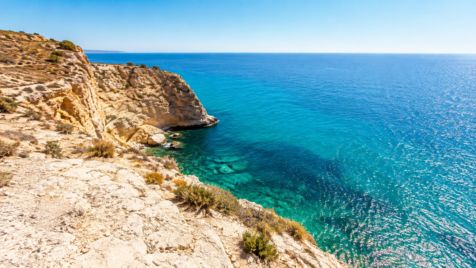 Côte méditerranéenne écrasée de soleil, eau turquoise et falaises sèches, surface de la mer vibrant sous la chaleur de midi.