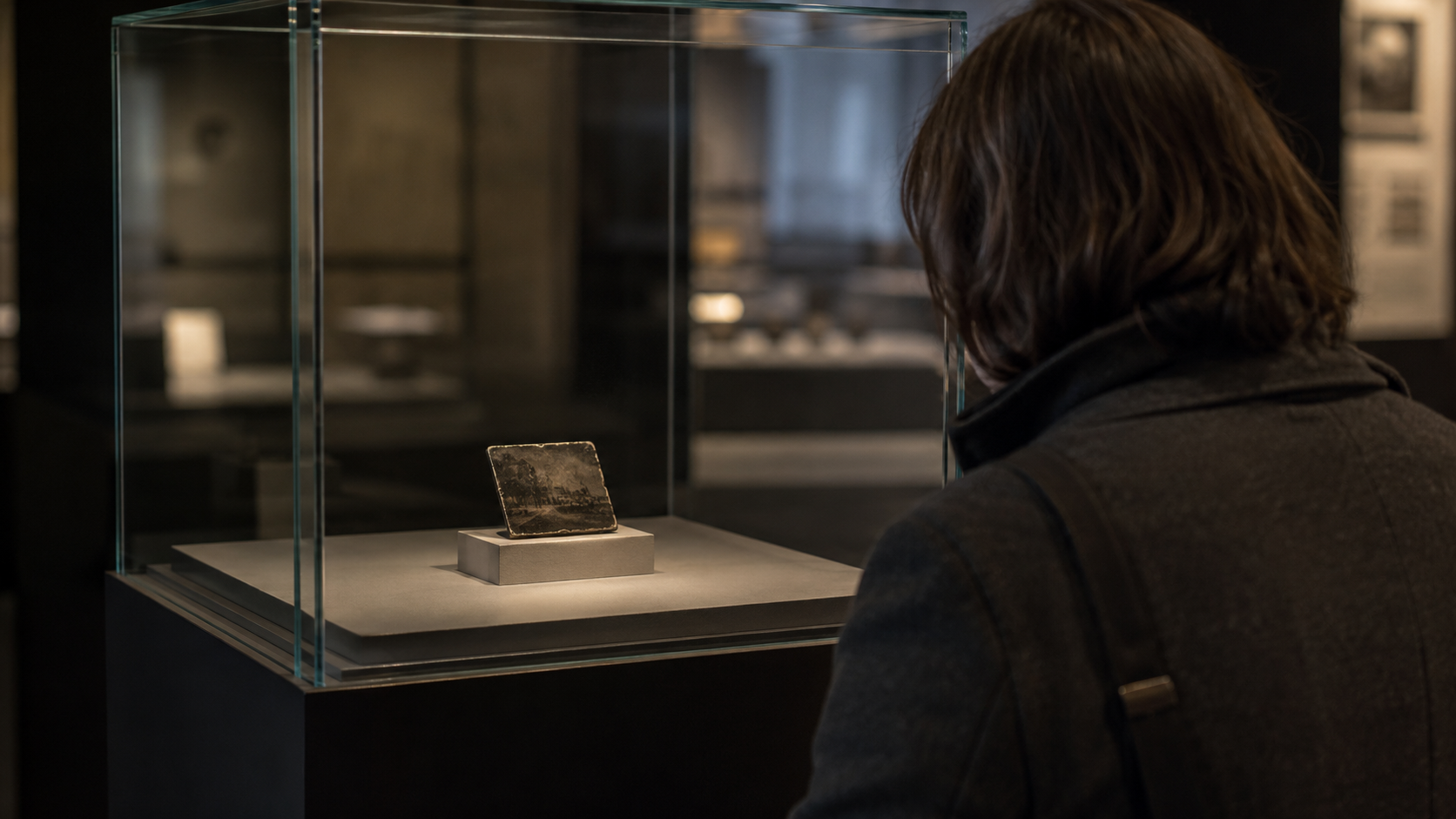 Petite plaque de métal terni exposée seule dans une vitrine fermée, un visiteur de dos penché vers le verre.
