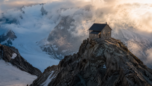 Petit refuge de haute montagne posé sur une arête rocheuse au-dessus d’un glacier, nuages bas en fin de journée.