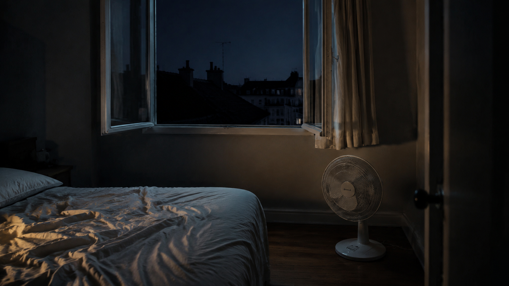 Fenêtre de chambre ouverte la nuit, rideau immobile et petit ventilateur posé au sol dans une pièce sombre.
