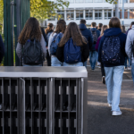 Lycéens vus de dos franchissant la grille d’un lycée français, casiers métalliques à téléphones au premier plan