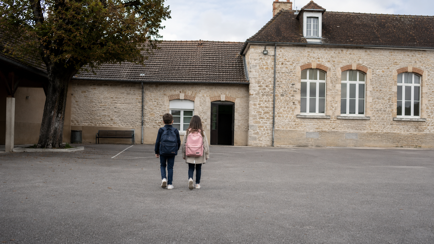 Petite école communale en pierre au bout d’une cour de récréation presque vide, deux enfants de dos marchant vers la porte.