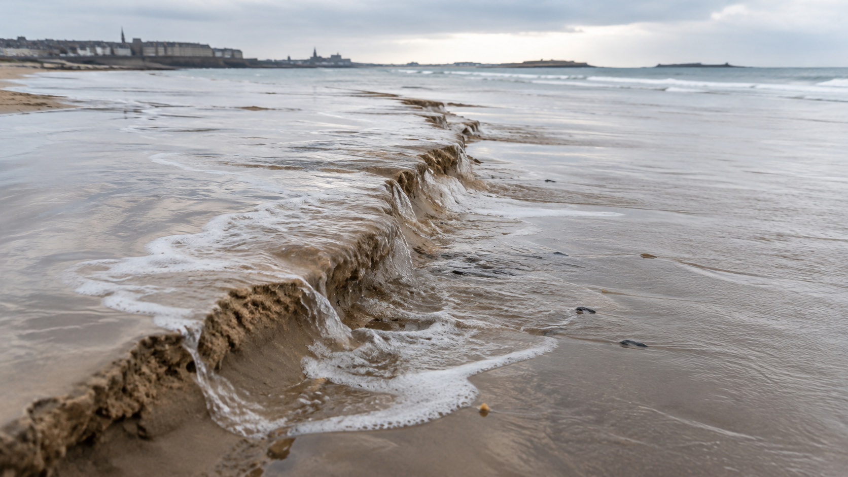 La mer montante recouvre une crête de sable humide qui s’effondre sur une plage large, écume étalée sur le muret aplati