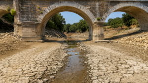 Lit de rivière asséché et craquelé sous un pont de pierre, un mince filet d’eau au centre.