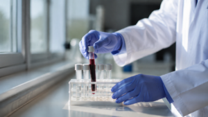 Des mains gantées posent un tube de prélèvement sanguin dans un portoir de laboratoire, près d’une fenêtre.