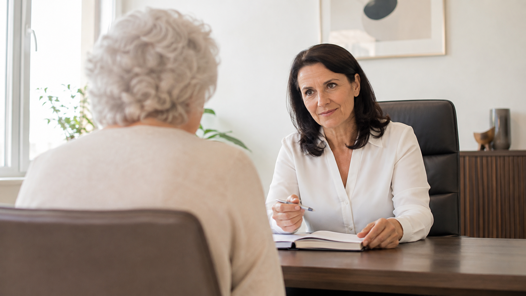 Une personne âgée, vue de dos, est assise face à un médecin dans un cabinet de consultation lumineux.