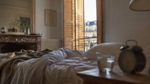 Chambre d’appartement français au petit matin, volets intérieurs en bois entrouverts, réveil analogique posé sur la table de nuit à côté d’un verre d’eau.