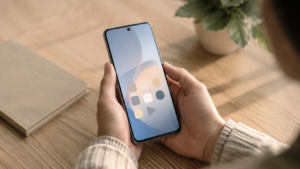 Deux mains tiennent un smartphone sans marque dont l'écran d'accueil n'affiche que quelques icônes neutres, au-dessus d'un bureau en bois clair.