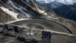 Route de col de haute montagne en épingle, roche nue et névés, chaises pliantes vides au bord de la chaussée