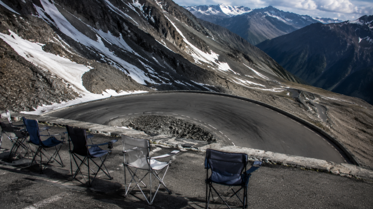 Route de col de haute montagne en épingle, roche nue et névés, chaises pliantes vides au bord de la chaussée