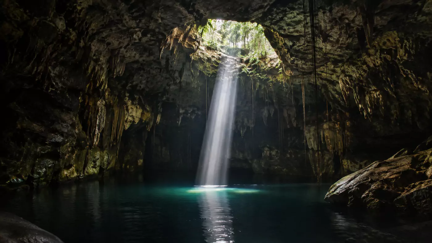 Cénote de jungle aux parois calcaires, eau sombre et immobile éclairée par un unique puits de lumière tombant du plafond