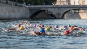 Nageurs en bonnets colorés serrés en peloton dans un fleuve urbain, quais de pierre et pont flous en arrière-plan.