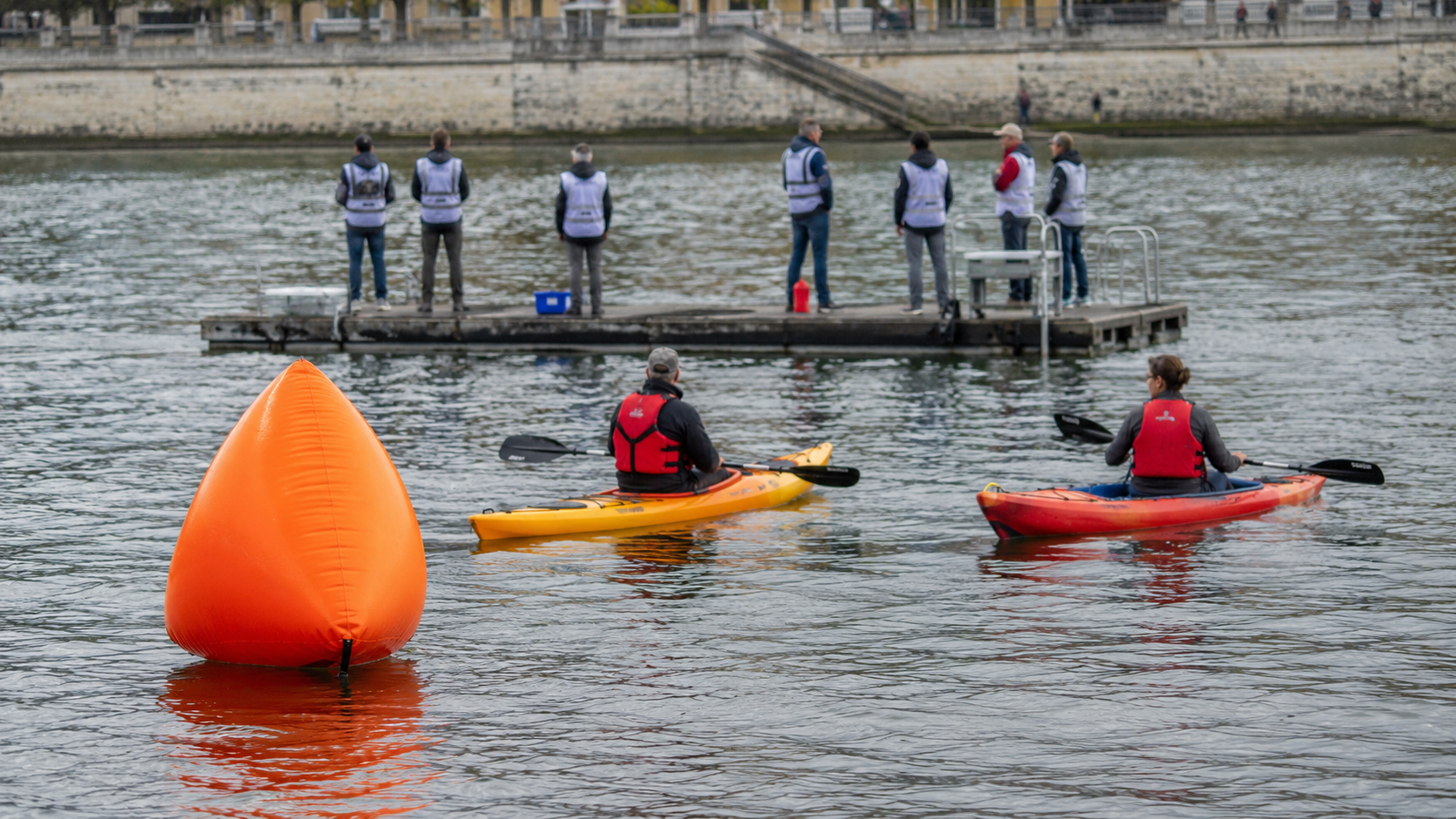 Une bouée de virage orange sur un fleuve, deux kayakistes de sécurité et des bénévoles en gilet observent depuis un ponton flottant.