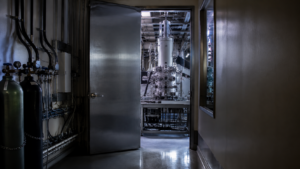 Couloir de laboratoire en fin de journée, porte métallique entrouverte laissant voir un grand instrument scientifique éclairé, câbles et bouteilles de gaz le long du mur, aucune personne visible