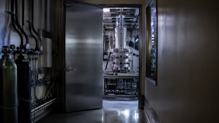 Couloir de laboratoire en fin de journée, porte métallique entrouverte laissant voir un grand instrument scientifique éclairé, câbles et bouteilles de gaz le long du mur, aucune personne visible