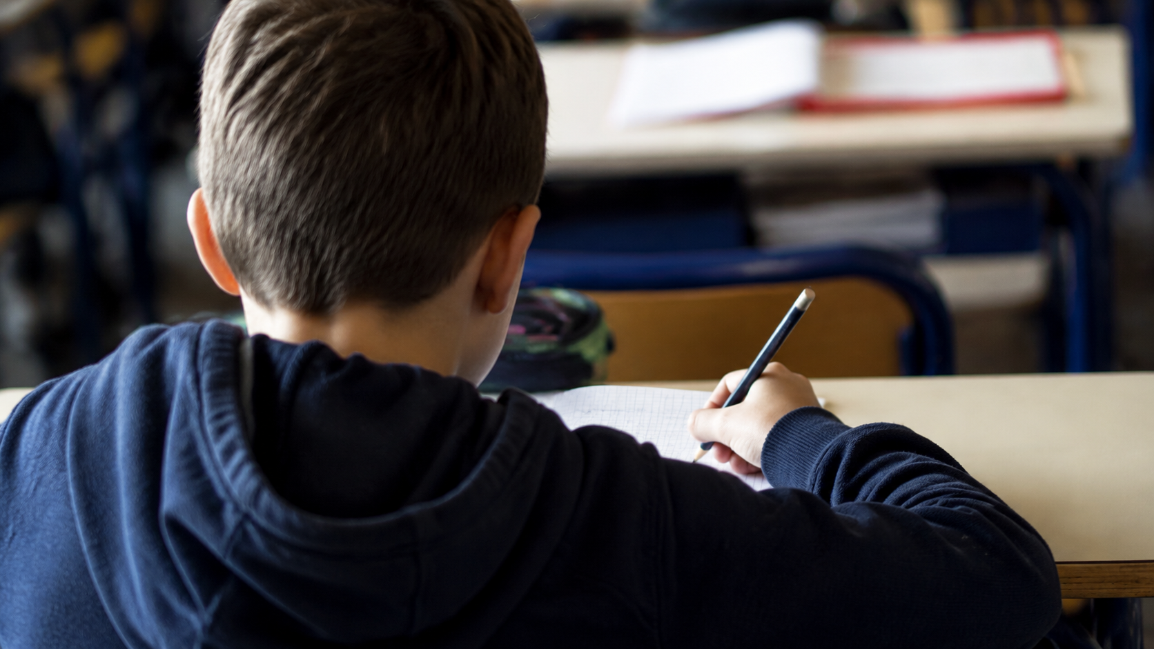 Élève vu de dos à son pupitre, stylo à la main, écrivant sur une feuille dans une salle de classe.