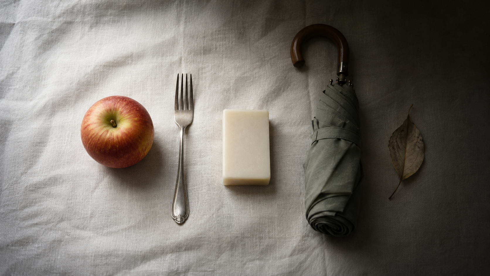 Objets domestiques du quotidien disposés à plat sur un lin clair et s'effaçant dans l'ombre : pomme, fourchette, savon, parapluie plié et feuille séchée.