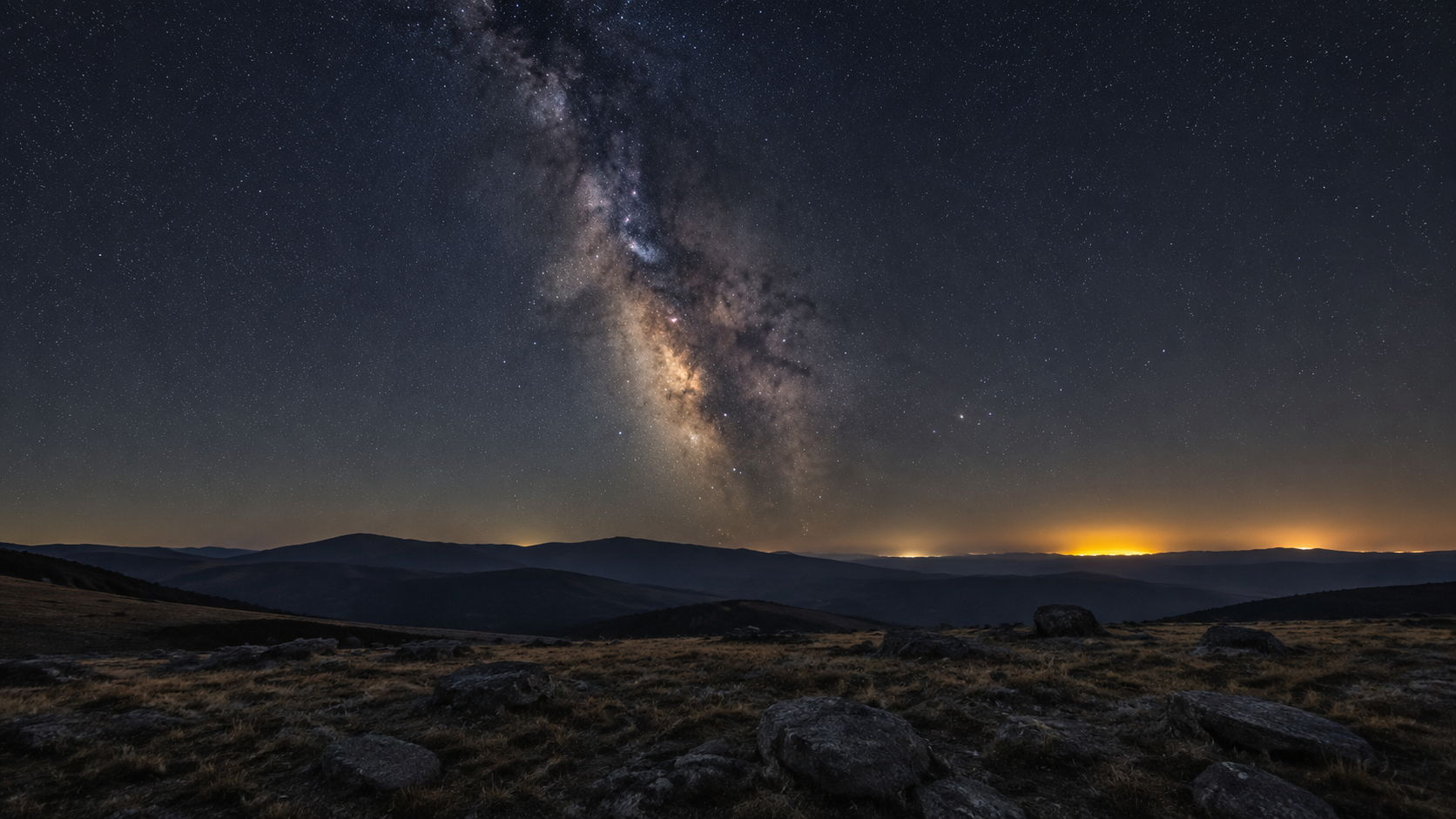 Paysage de hauts plateaux la nuit, la Voie lactée nettement visible au-dessus des collines, avec une lueur artificielle lointaine sur l’horizon.