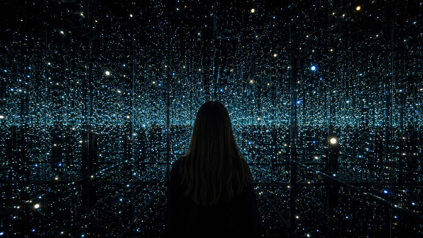 Un visiteur de dos dans une salle obscure tapissée de miroirs où des points lumineux se répètent à l’infini.