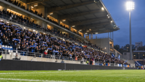 Tribune latérale d’un stade de football français aux deux tiers garnie, écharpes levées, en début de soirée sous les projecteurs.