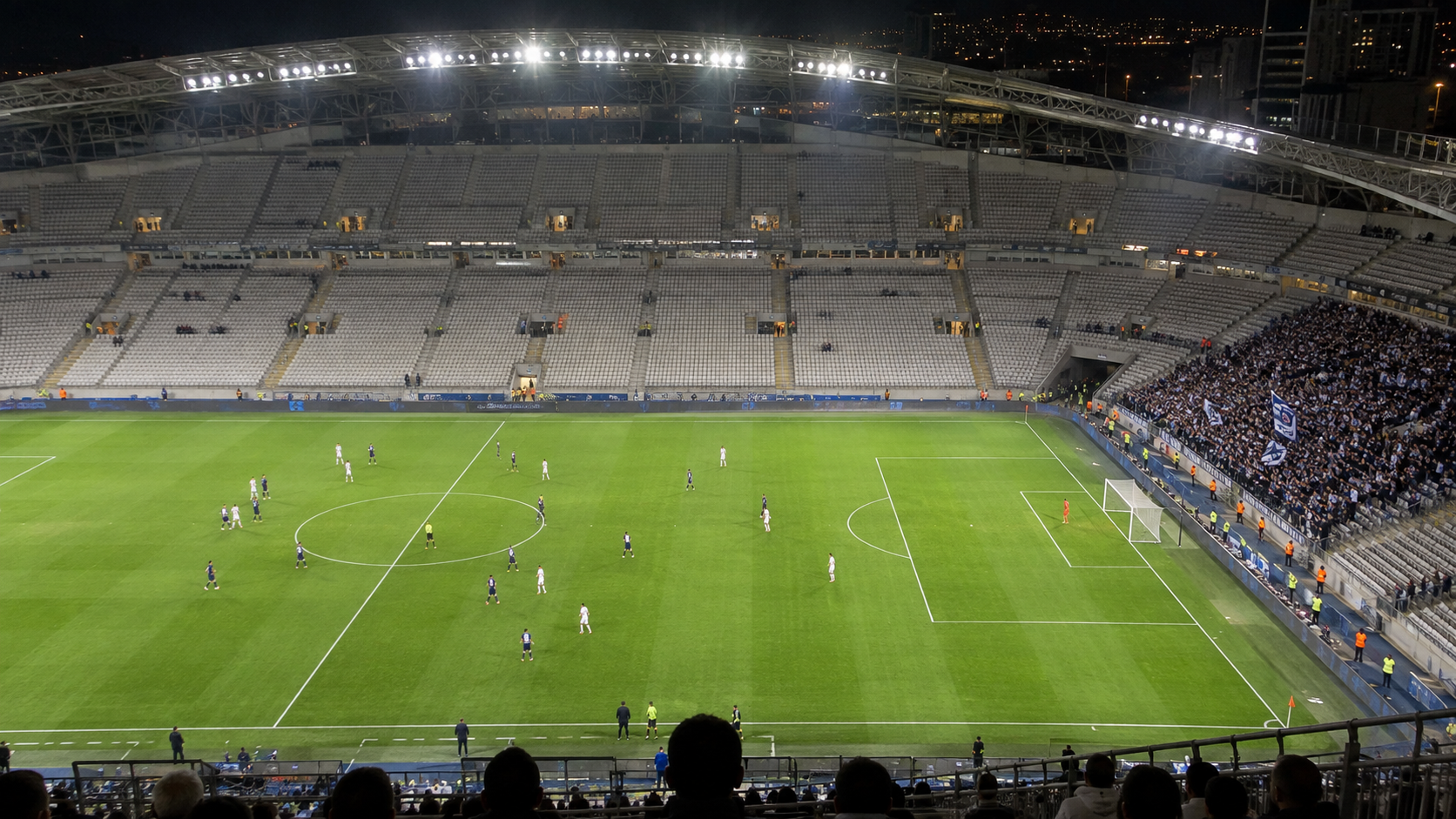 Vue d’un stade de football depuis les gradins hauts, une tribune presque vide face à un parcage visiteurs dense et debout.