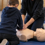 Les mains d’un enfant posées sur le thorax d’un mannequin de réanimation, guidées par celles d’un adulte, dans un gymnase scolaire.