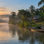 Village amazonien au bord d’une rivière à l’aube, maisons de bois à toit de palmes et pirogue amarrée sur la berge.