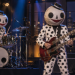 Deux musiciens en costumes à pois et têtes de papier mâché sur une scène de plateau télé, batterie à gauche, guitare double manche à droite.