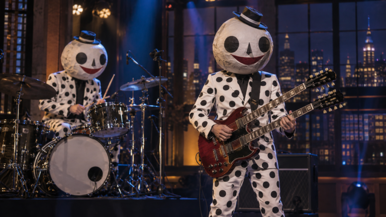Deux musiciens en costumes à pois et têtes de papier mâché sur une scène de plateau télé, batterie à gauche, guitare double manche à droite.