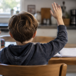 Un enfant de dos, assis à la table de la cuisine, lève la main au-dessus d'un cahier ouvert.