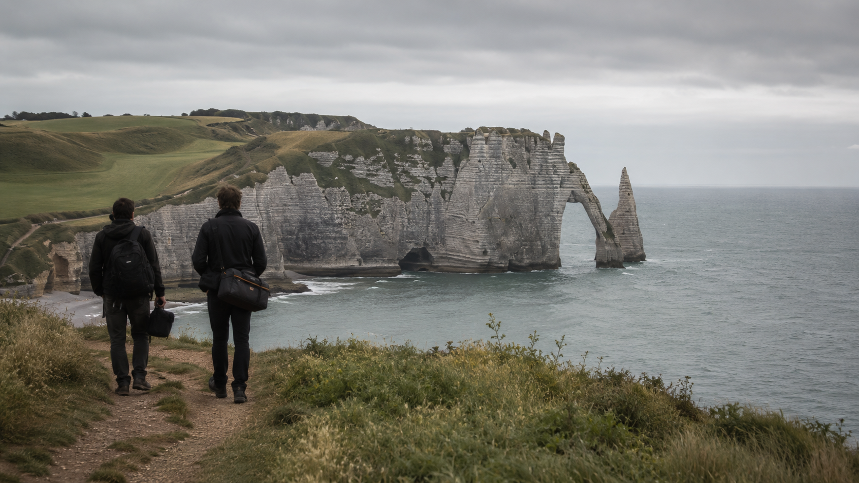 Les falaises de craie et l’arche naturelle d’Étretat, en Normandie, sous une lumière couverte, avec deux silhouettes de dos portant un sac de caméra.