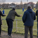 Des adultes debout derrière la main courante d’un terrain de football de jeunes, vus de dos.