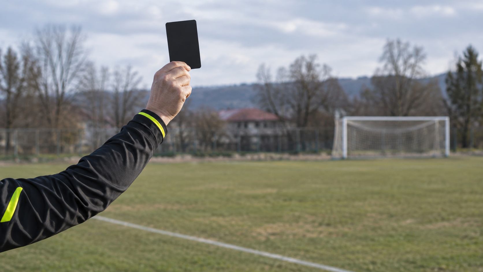 La main d’un arbitre lève un carton noir au bord d’un terrain de jeunes, sans visage visible.