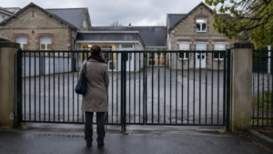 Portail fermé d’une école primaire française un matin gris, un adulte de dos devant la grille.