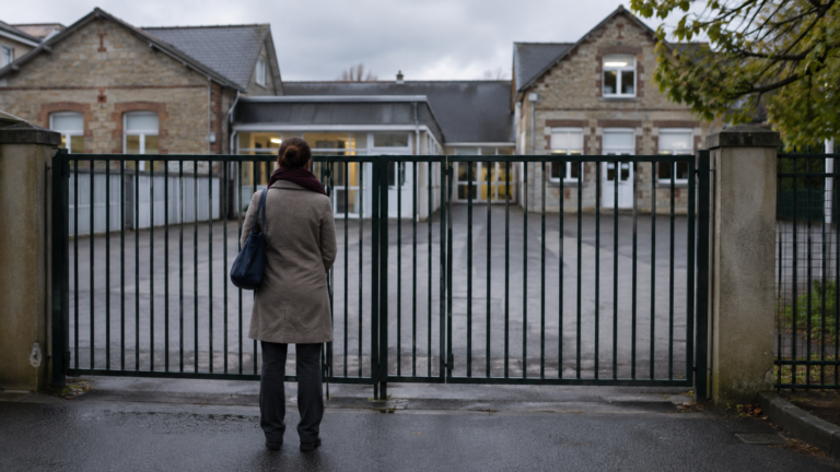 Portail fermé d’une école primaire française un matin gris, un adulte de dos devant la grille.