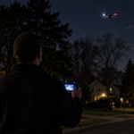 De dos, un homme tient une télécommande de drone éclairée dans une rue pavillonnaire de nuit, un quadricoptère en vol au-dessus des arbres sombres.
