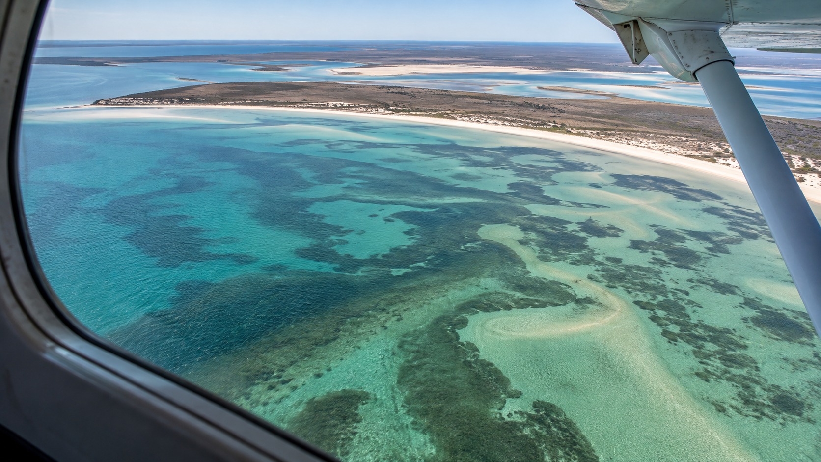 Vue plongeante depuis le hublot d’un petit avion sur une baie peu profonde, bancs de sable turquoise et herbiers sombres, hauban d’aile dans le cadre.