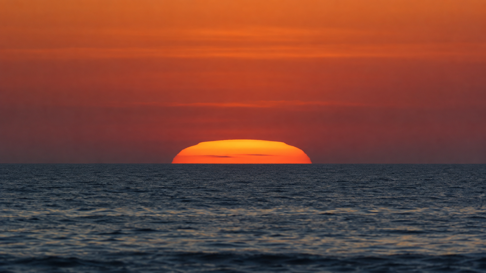 Disque solaire orange aplati par la réfraction, à moitié sous un horizon marin dégagé