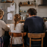 Quatre personnes vues de dos autour d’une table de cuisine française, un cartable ouvert sur une chaise et des papiers administratifs aimantés sur le frigo.