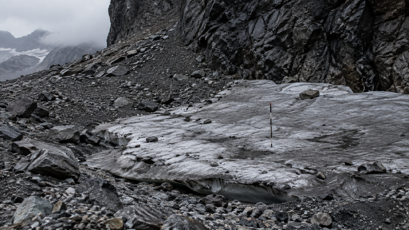 Une petite plaque de glace grisâtre de quelques dizaines de mètres, coincée au pied d’une paroi rocheuse sombre au milieu des éboulis, avec une perche graduée plantée dedans.