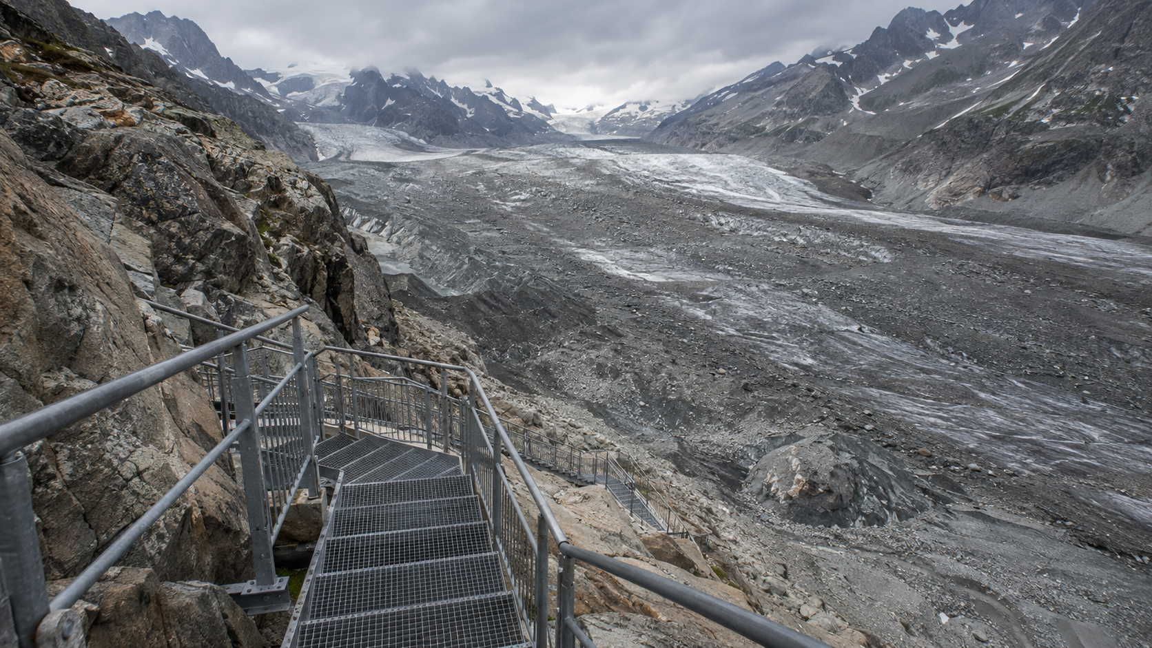 Un long escalier métallique fixé à une paroi rocheuse descend vers une large langue glaciaire couverte de débris, sous un ciel gris.
