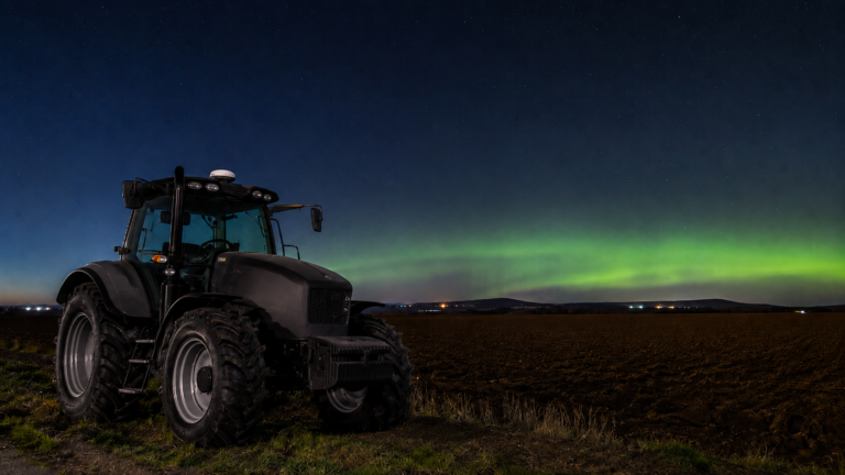 Tracteur à l’arrêt en bordure d’un champ labouré la nuit, petit récepteur satellite sur le toit de la cabine, faible lueur verte à l’horizon.