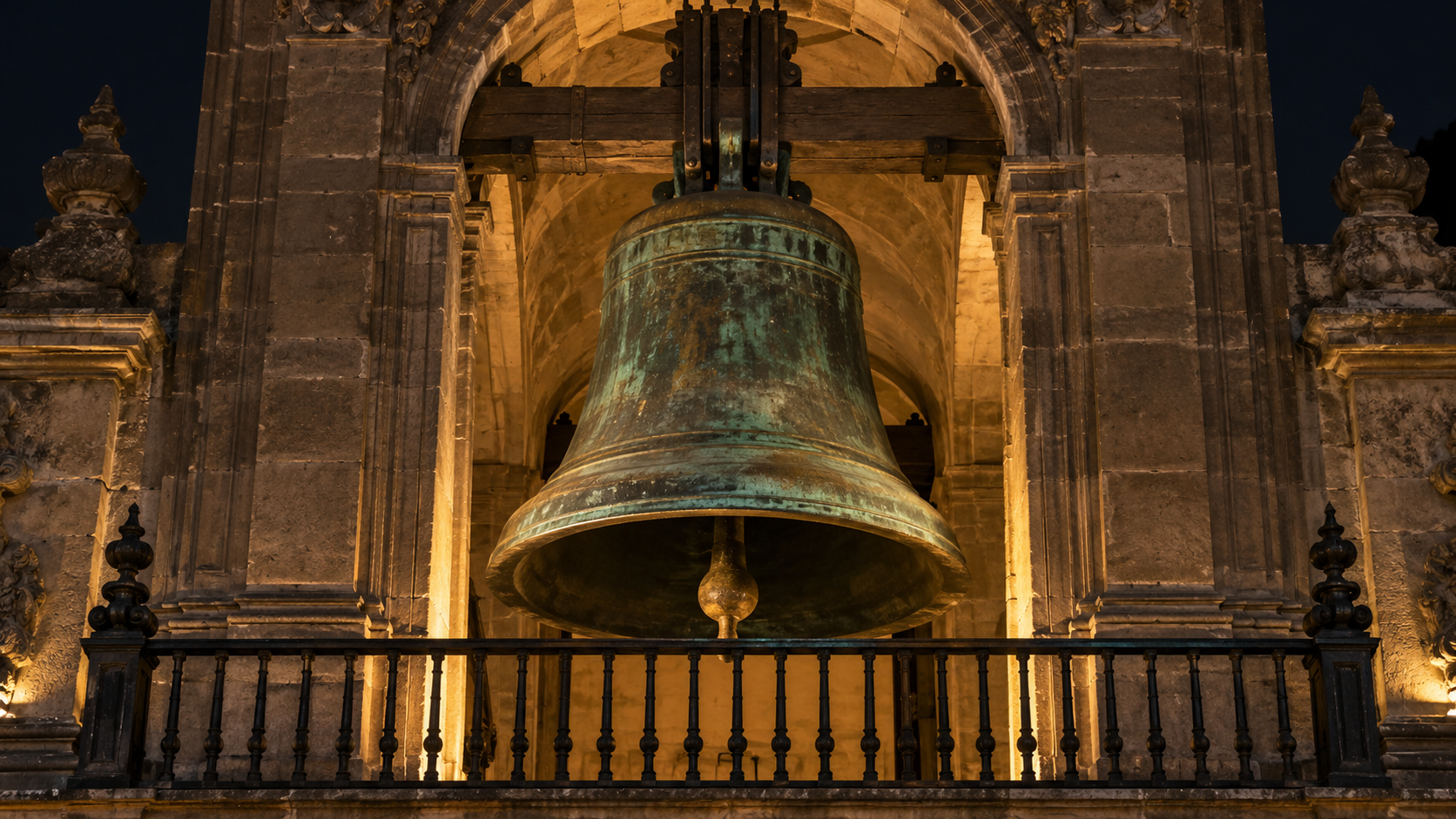 Grande cloche de bronze patiné installée dans une niche de pierre au-dessus d’un balcon de palais, éclairée la nuit.